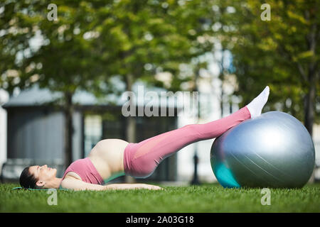 Jeune femme enceinte dans un vêtement de sport rose couché sur tapis d'exercice et l'entraînement avec fitness ball au cours de l'entraînement sportif dans le parc Banque D'Images