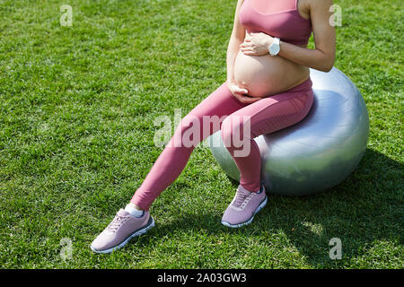 Close-up of young pregnant woman in pink clothing sitting on fitness ball toucher son ventre et faire exercice de relaxation en plein air Banque D'Images