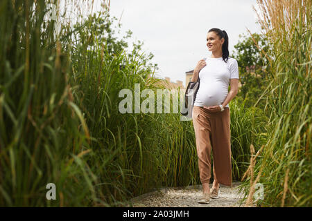 Young pregnant woman in casual clothing marcher le long de la haute vert et profiter de la nature plantes Banque D'Images