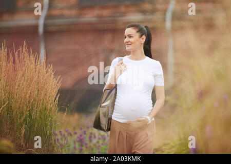 Jeune femme enceinte aux cheveux noirs portant des vêtements décontractés marcher dans la ville et souriant Banque D'Images