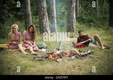 Les filles assis près d'un feu de camp alors que la lecture de livres. Barbu allongé sur l'herbe dans la forêt. Amis camping en été Banque D'Images