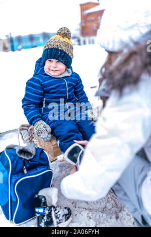 Petit garçon de 3,5 ans en salopette bleue, en hiver sur une patinoire de la ville, est assis sur un banc, mère d'un enfant chaussures femme, chaussures, patins, week-end Banque D'Images