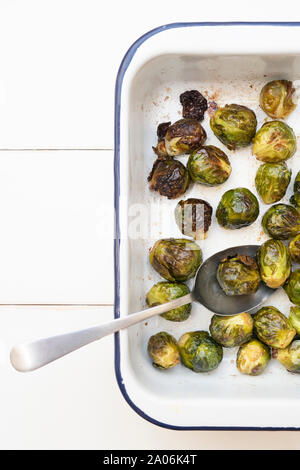 Brassica oleracea. Choux de Bruxelles rôtis dans un plat de cuisson en émail blanc avec une cuillère sur fond de bois blanc Banque D'Images