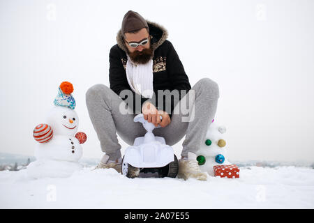 Hipster équitation petite voiture sur ciel blanc. Homme barbu à lunettes pilote et un chapeau. Bonhomme de neige et l'arbre de Noël sur paysage d'hiver. Maison de vacances concept célébration Banque D'Images