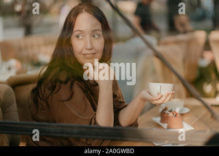 Charmante femme brune avec de longs cheveux bouclés assis à fenêtre dans café avec tasse de café dans les mains Banque D'Images