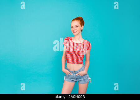 Belle positive attractive cheerful skinny fille rousse avec bun, vêtus de t-shirt et un short, tenant les mains dans les poches, isolé sur teal bri pastel Banque D'Images