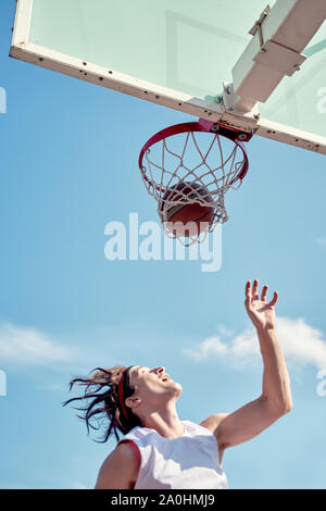 Photo de sports man throwing ballon dans panier de basket-ball sur le terrain de sport de rue contre le ciel bleu Banque D'Images