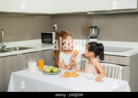 Jolie jeune femme et sa fille peu cute mangent des gâteaux et des cookies sur cuisine et drinking orange Banque D'Images