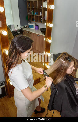 Coiffeur professionnel girl crêper les cheveux bruns longs avec des fers de cheveux. Processus de la coiffure en salon de beauté Banque D'Images