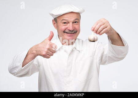 Hispanic male mature chef cuisinier en uniforme blanc et l'ail holding showing thumb up. Bon pour l'hiver d'épices repas. Banque D'Images