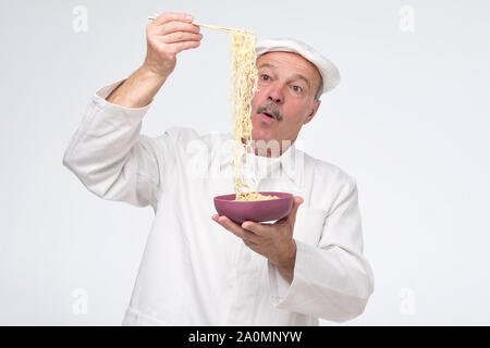 Chef cuisinier Senior hispanic man eating nouilles asiatiques tenant une longue mèche dans sa main. Studio shot Banque D'Images