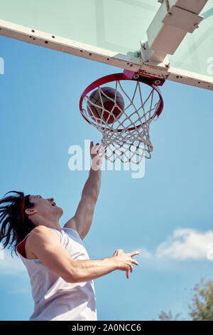 Image de l'athlète man throwing ballon dans panier de basket-ball sur le terrain de sport de rue contre le ciel bleu Banque D'Images