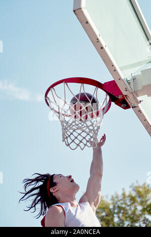 Photo de jeune athlète man throwing ballon dans panier de basket-ball sur le terrain de sport de rue contre le ciel bleu Banque D'Images