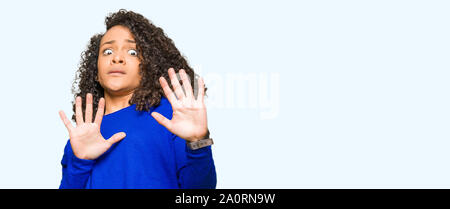 Belle jeune femme aux cheveux bouclés wearing winter sweater peur et terrifié par la peur d'arrêt expression geste avec les mains, criant sous le choc. Pan Banque D'Images