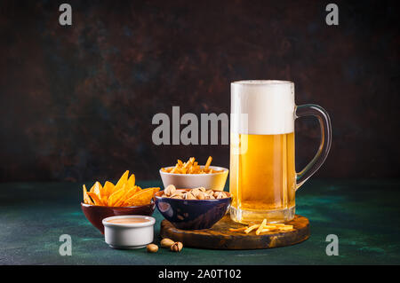 Mug en verre avec de la bière avec de la mousse et l'eau et des en-cas sur un fond vert foncé Banque D'Images