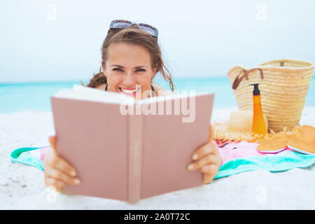 Happy trendy 40 ans femme aux longs cheveux bouclés en noir élégant maillot sur une plage blanche livre de lecture. Banque D'Images