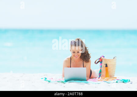 Heureux 40 ans élégante femme aux longs cheveux bouclés dans un élégant maillot noir sur une plage de sable blanc à l'aide d'internet sur un ordinateur portable. Banque D'Images