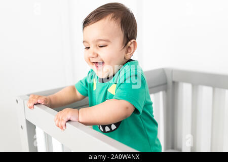 Bébé debout et se tenant à la rambarde du lit. Portant un t-shirt vert mignon dans une pépinière moderne et minimaliste lumineux chambre à coucher. Banque D'Images