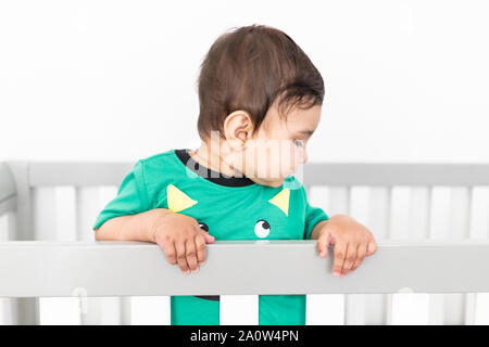 Bébé debout et se tenant à la rambarde du lit. Portant un t-shirt vert mignon dans une pépinière moderne et minimaliste lumineux chambre à coucher. Banque D'Images