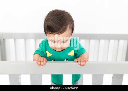 Bébé debout et se tenant à la rambarde du lit. Portant un t-shirt vert mignon dans une pépinière moderne et minimaliste lumineux chambre à coucher. Banque D'Images