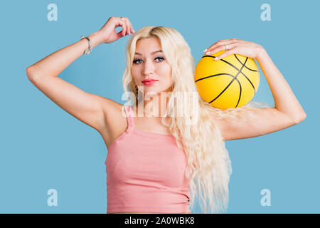 Jeune femme séduisante avec de longs cheveux blonds, beau maquillage, manucure portant haut court rose holding basketball looking at camera with pleasant expressi Banque D'Images