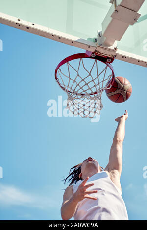 Photo d'athlète man throwing ballon dans panier de basket-ball sur le terrain de sport de rue contre le ciel bleu Banque D'Images