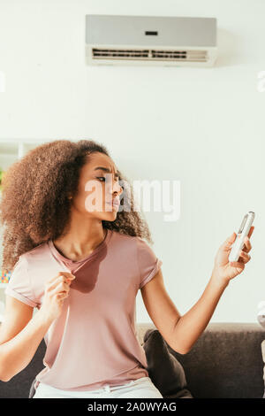 Insatisfait african american woman in t-shirt en sueur à thermomètre à tout en souffrant de la chaleur à la maison Banque D'Images