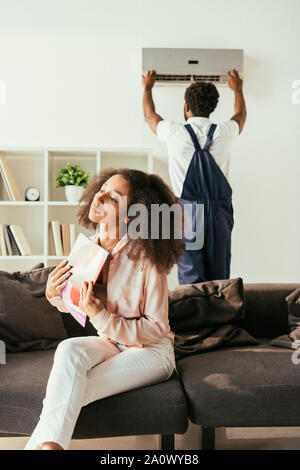 Young african american woman sitting on sofa et forme avec magazine, tandis que l'Amérique africaine climatiseur fixation réparateur Banque D'Images