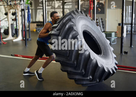 Vue latérale sur toute la longueur de l'homme afro-américain musculaire flipping tire au cours de l'exercice dans la salle de sport moderne, copy space Banque D'Images