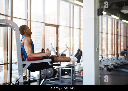 Vue de côté portrait de l'homme afro-américain musculaire à l'aide de machines d'exercice lors de l'entraînement dans la salle de sport moderne contre la fenêtre, copy space Banque D'Images