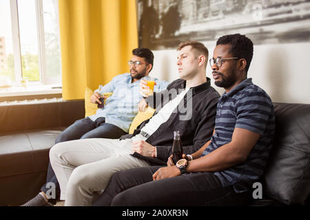 L'amitié, sports et divertissements concept. Vue de côté de trois amis de regarder la télévision la consommation de boissons gazeuses et de jus à la maison wit Banque D'Images