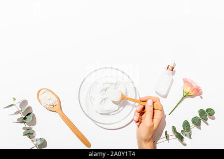 Woman's hand holding spoon d'argile cosmétique de faire masque de visage à côté de bouteille de branches d'eucalyptus et de sérum sur fond blanc avec copie espace, Fla. Banque D'Images