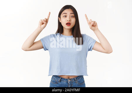 Surpris se demanda cute asian jeune adolescente curieuse bouche ouverte mise en doute soulever index haut publicité pointant vers le haut, regarder comme intrigué l'appareil photo Banque D'Images