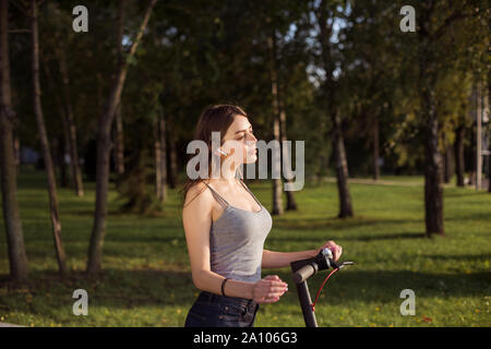 Brunette girl riding un éco-friendly electric scooter de coup dans un parc par temps ensoleillé sur les trottoirs Banque D'Images