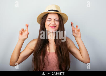 Heureux happy woman with closed eyes strabisme en t-shirt rose en priant avec les doigts croisés sur fond bleu. Portrait Banque D'Images