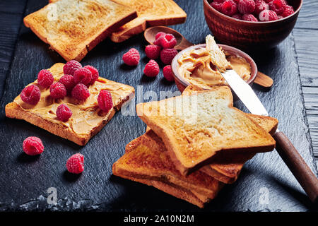 Close-up of open sandwiches de pain grillé avec du beurre d'arachide et de framboises fraîches sur un plateau en ardoise noire sur une table de cuisine en bois noir, hor Banque D'Images