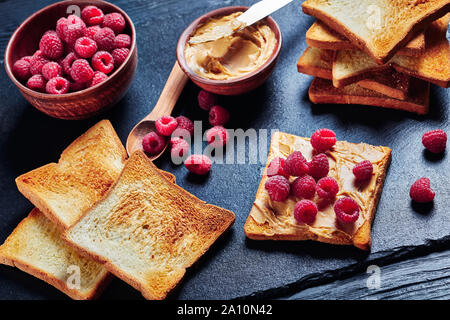 Close-up de toasts avec de doux le beurre d'arachide et de framboises fraîches sur un plateau en ardoise noire sur une table de cuisine en bois noir, vue horizontale à partir de ci-dessus, f Banque D'Images