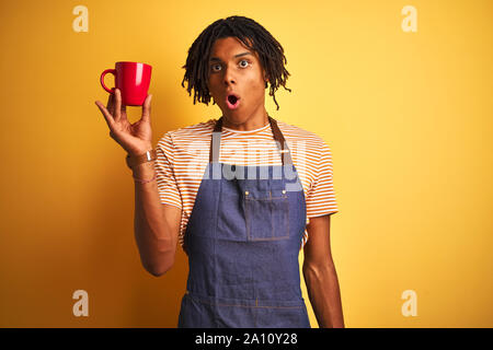 L'homme avec des dreadlocks Afro barista tasse de café potable sur fond jaune isolé peur sous le choc avec un visage surpris, effrayé et excité avec Banque D'Images