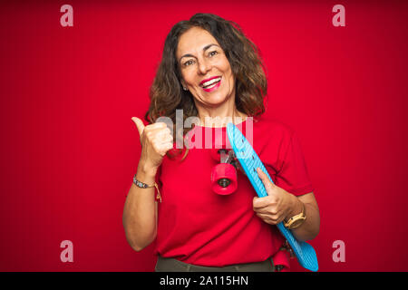 L'âge moyen du patineur senior woman holding skateboard rouge sur fond isolé et de pointage montrant avec le pouce jusqu'à la côte avec happy face smiling Banque D'Images