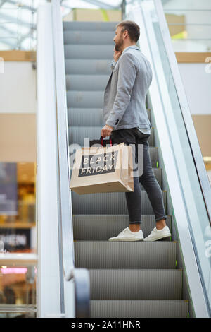 Vue arrière de la mode portrait man holding shopping bags avec inscription Vendredi Noir debout sur escalator dans mall, copy space Banque D'Images