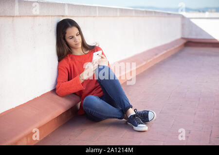 Chica adolescente mirando el sentada smartphone en la azotea Banque D'Images