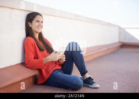 Chica adolescente sonriente mirando el sentada smartphone en la azotea Banque D'Images