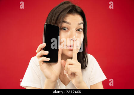 Portrait of young woman's portrait rouge sur fond de studio. Beau modèle féminin en chemise blanche. Concept d'émotions humaines, expression du visage, les ventes. Holding phone, whispering a secret. Banque D'Images