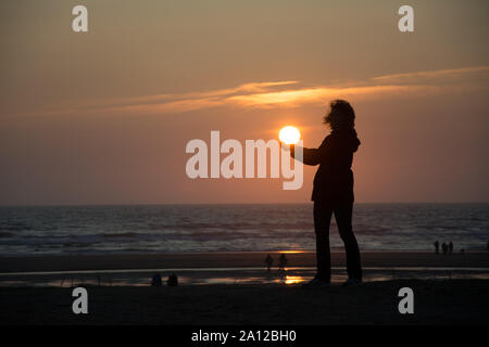 Silhouette d'une femme debout sur une plage de l'océan, tenant dans ses mains un coucher de soleil Banque D'Images