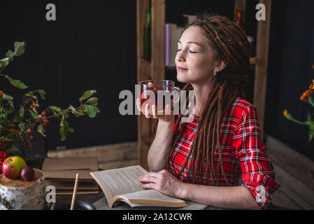 Belle femme lisant un livre romantique dans une atmosphère chaleureuse et confortable profitant de l'ambiance d'automne. Table rustique avec des fleurs jaunes et une pile de livres Banque D'Images