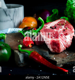 Steak juteux se trouve sur une pierre sauvage à côté de piments rouges et verts, des oignons et un bleu salade verte fraîche sur une table en bois noir Banque D'Images