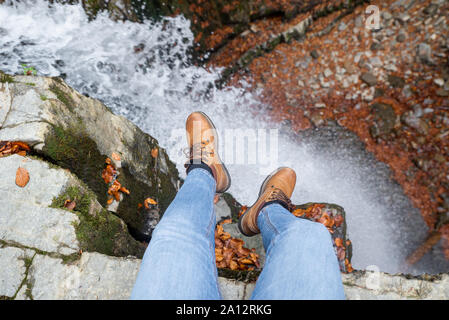 Les jambes de l'homme en jeans et bottes marron assise sur le bord à la cascade à Banque D'Images