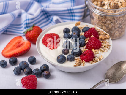 Le yogourt dans un bol blanc avec le gruau et les fraises, bleuets et framboises sur le dessus. Banque D'Images