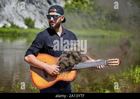 Dans les mains du guitariste guitare, où il y a de la fumée. Un homme barbu d'une casquette, lunettes de soleil, et une chemise noire joue une guitare acoustique, d'où t Banque D'Images