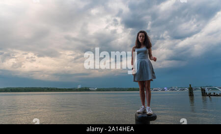 Teen girl in dress debout sur bollard en face de la rivière Potomac, avec un téléphone mobile dans sa main Banque D'Images